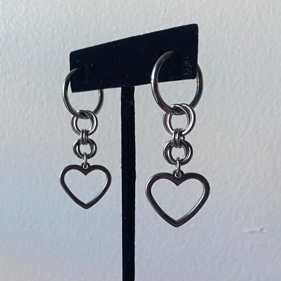 Stainless steel huggie hoop earrings with two rows of interlocking jump rings descending and an open heart charm hanging from a black velvet earring stand.