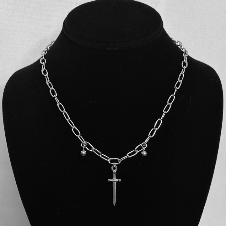 A necklace with stainless steel paperclip chain with a dagger charm at the center and onw small stainless steel ball charm on either side displayed on a black velvet neckform.